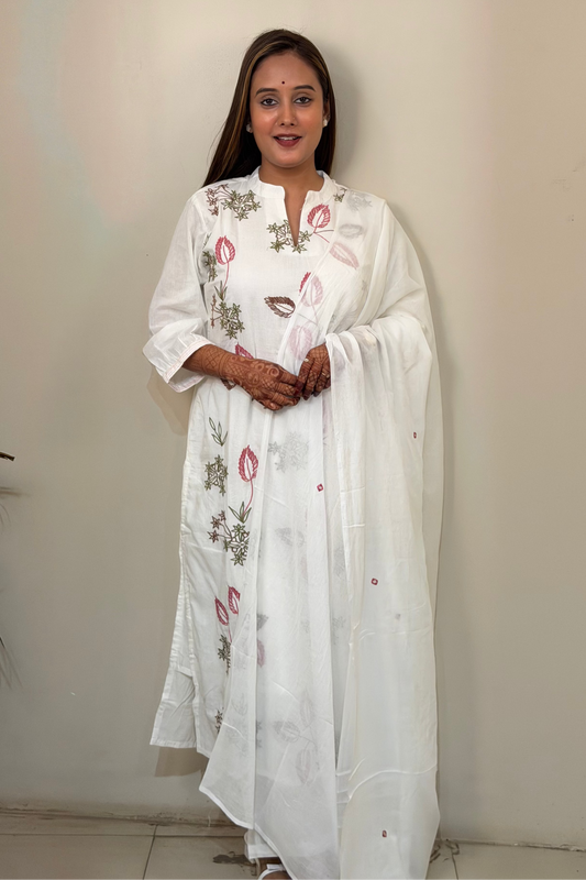 Woman wearing a white embroidered dress against a plain wall.